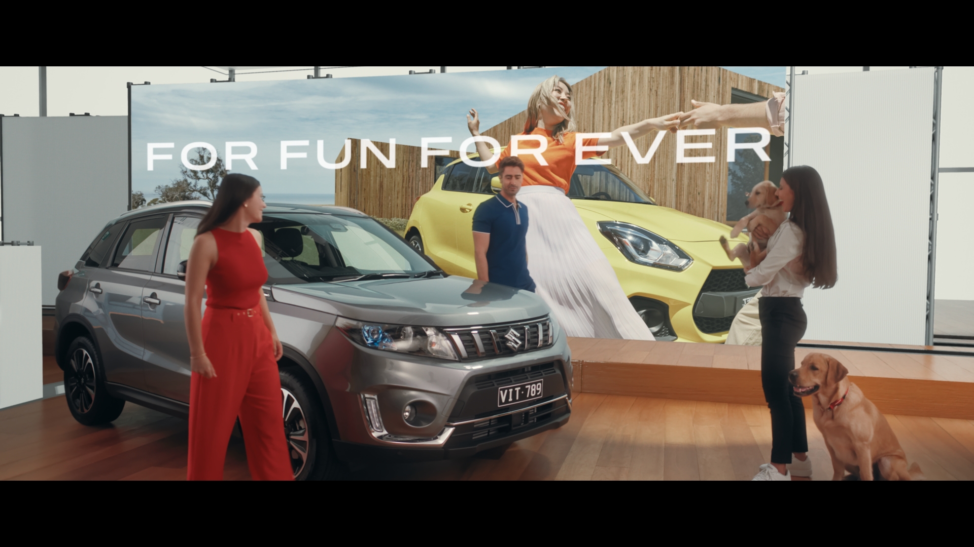 Two women, a man, and two dogs are in a studio with two Suzuki cars and a billboard promoting 'Fun Forever'.