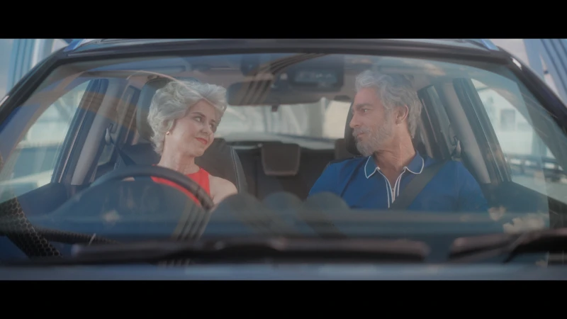 Mature couple in blue car, driving on bridge. Woman in red dress smiles at man in driver's seat with urban backdrop.