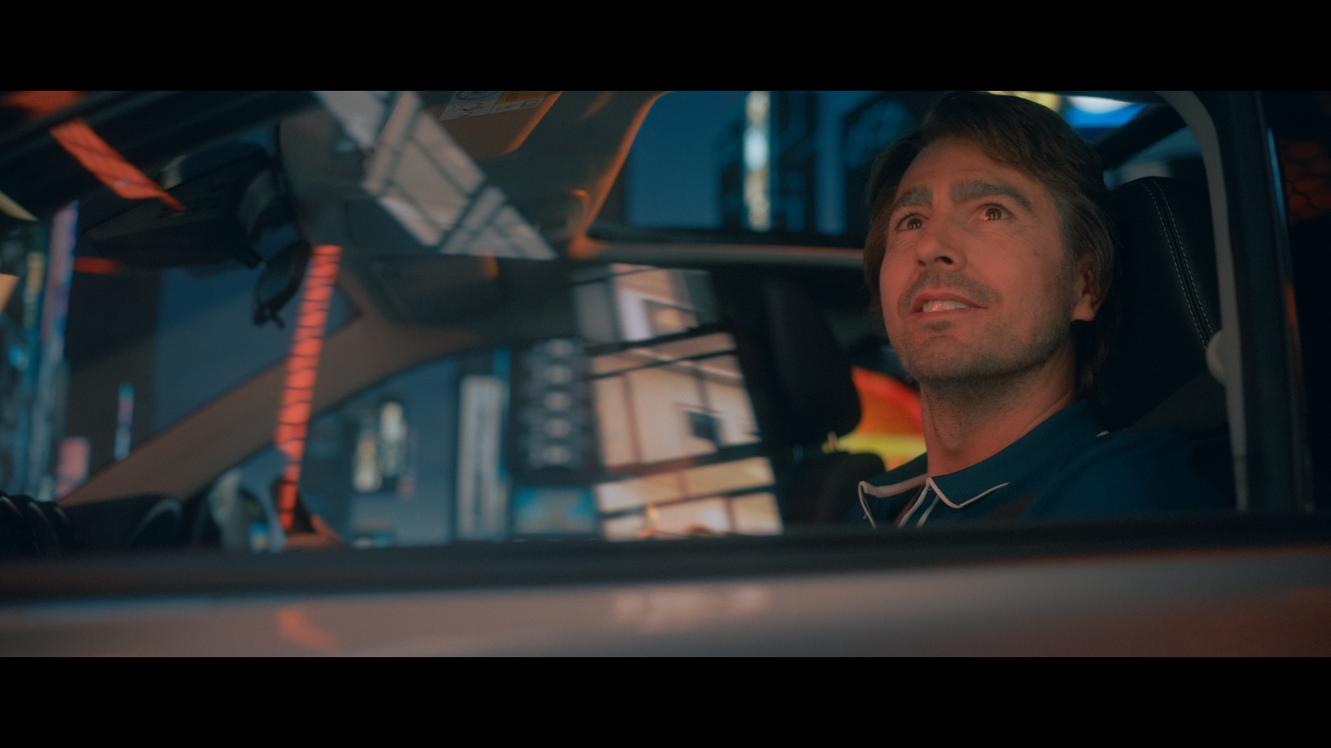 Adult male in a car at night, cityscape background with colorful lights, captured in medium shot showing a contemplative mood.