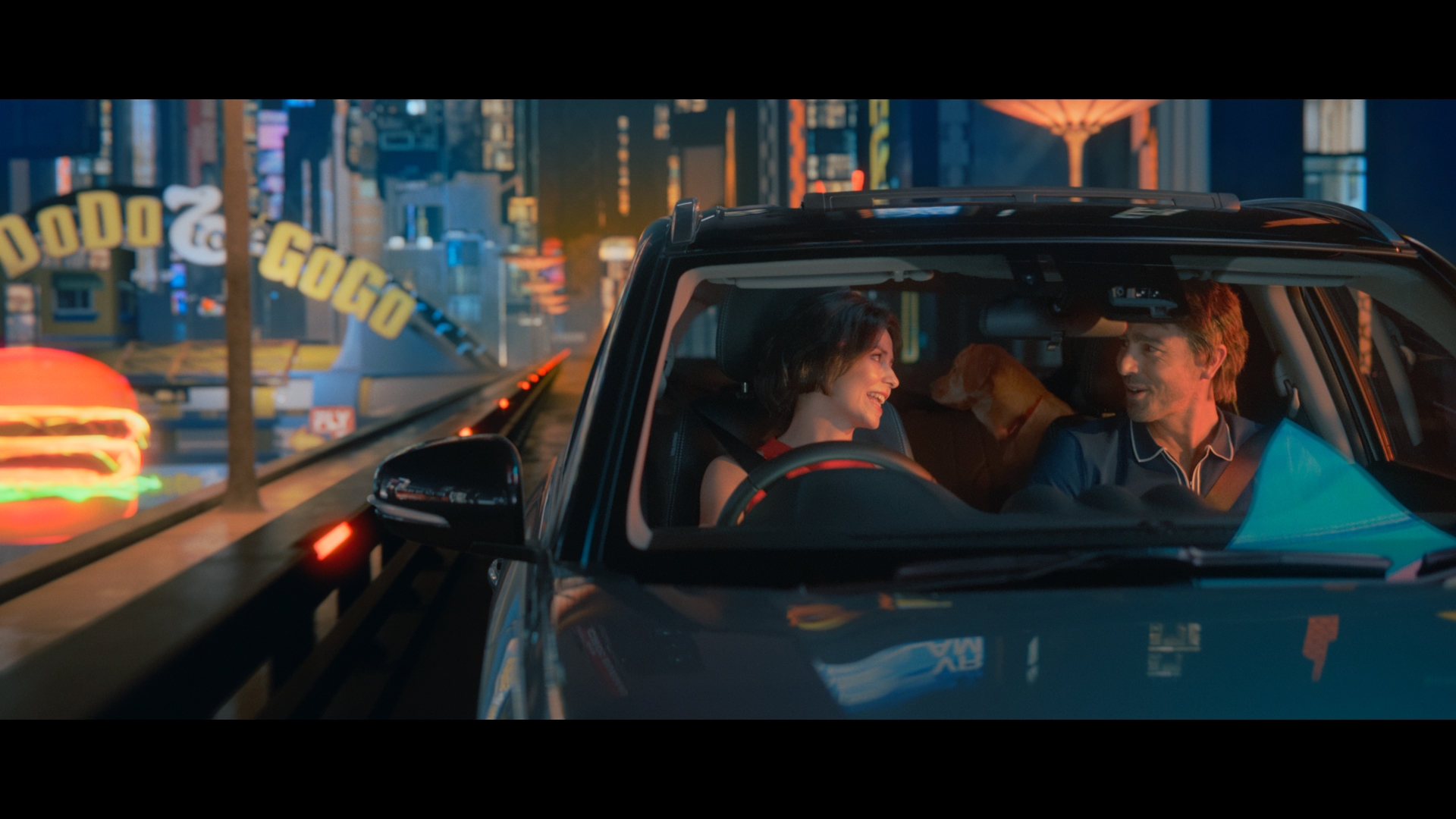 Adult male and female couple with a dog sit inside an SUV while driving at night in an urban environment with bright neon signs.