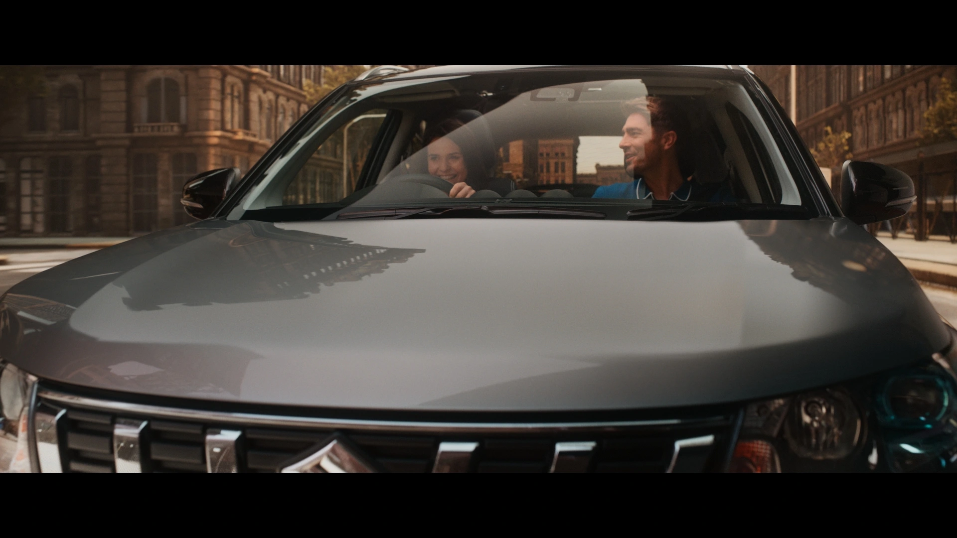 Front view of a silver Suzuki SUV on a city street with a smiling couple sitting inside.