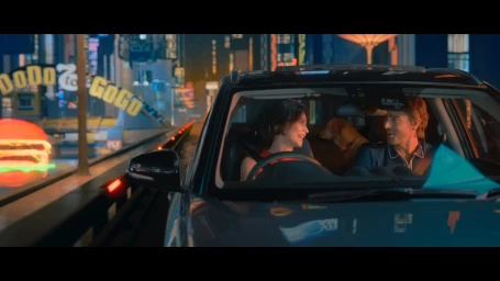 Man and woman smiling inside a car driving through a neon-lit city at night with a dog in the back seat