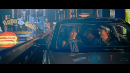 Woman in red top driving a car while talking to a passenger, viewed from the backseat.