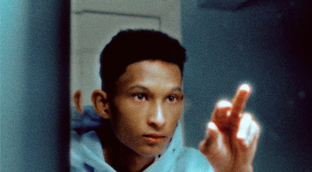 A young man in a light blue hoodie reaches toward a reflective surface with a glowing finger in a cool-toned room.
