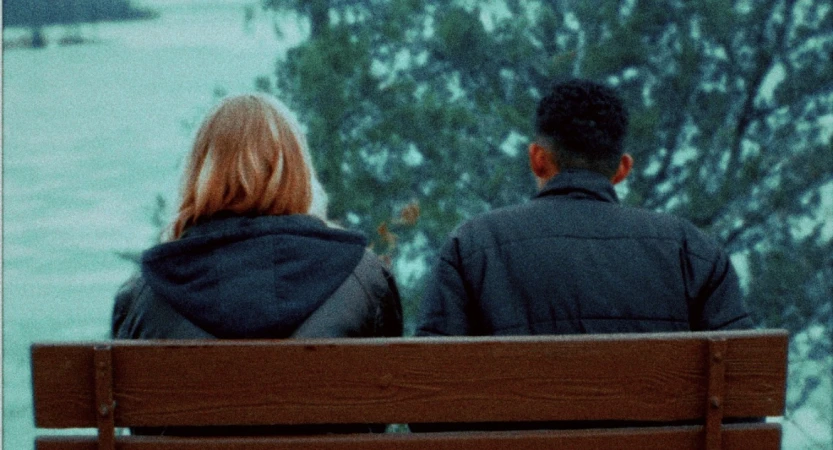 Two people seated side by side on a wooden bench viewed from behind, looking out towards water and lush green trees.