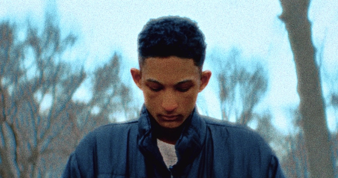 Young man looking downward outdoors wearing a dark jacket with bare trees and sky in the background.