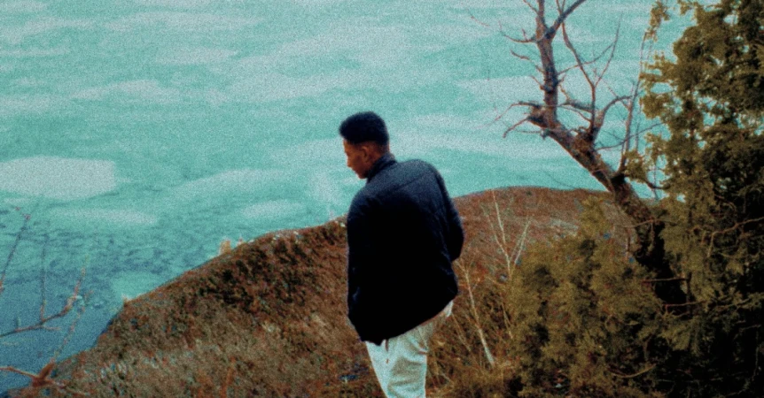 Man in a dark jacket looking down from a hillside over bright turquoise water.