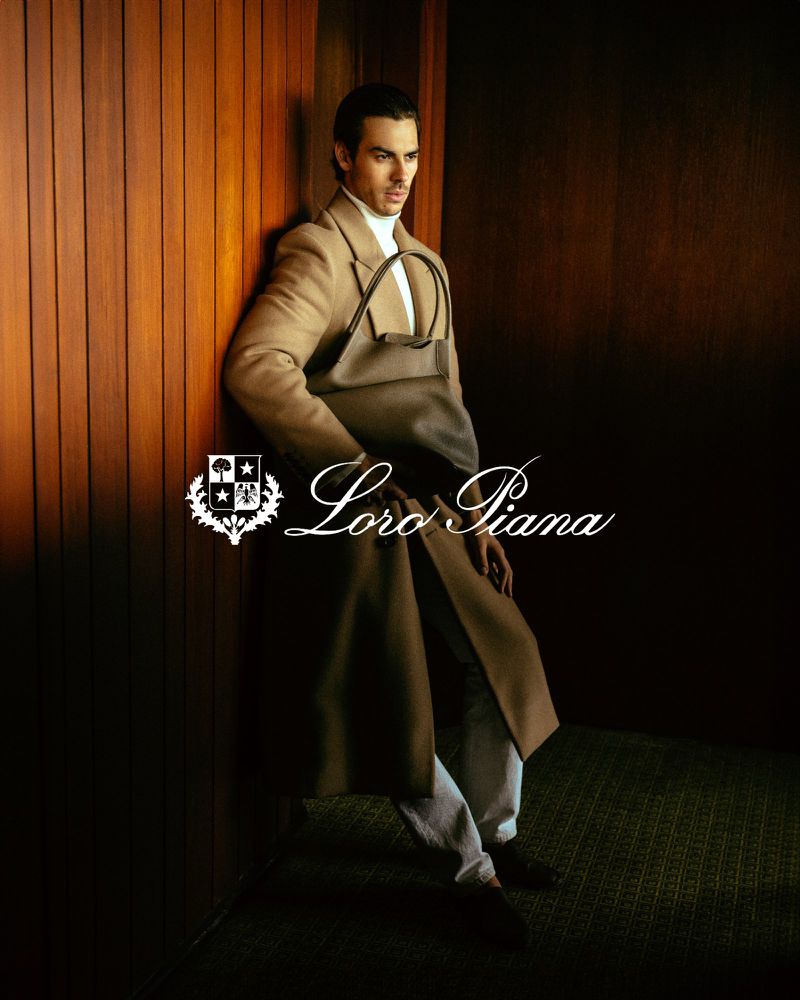 Man in a tan coat and turtleneck leaning against a paneled wall with a handbag in an indoor setting.