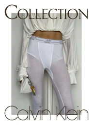 A model wearing sheer white Calvin Klein branded leggings and a gathered cream top, holding a small pouch with keys against a pale wall.