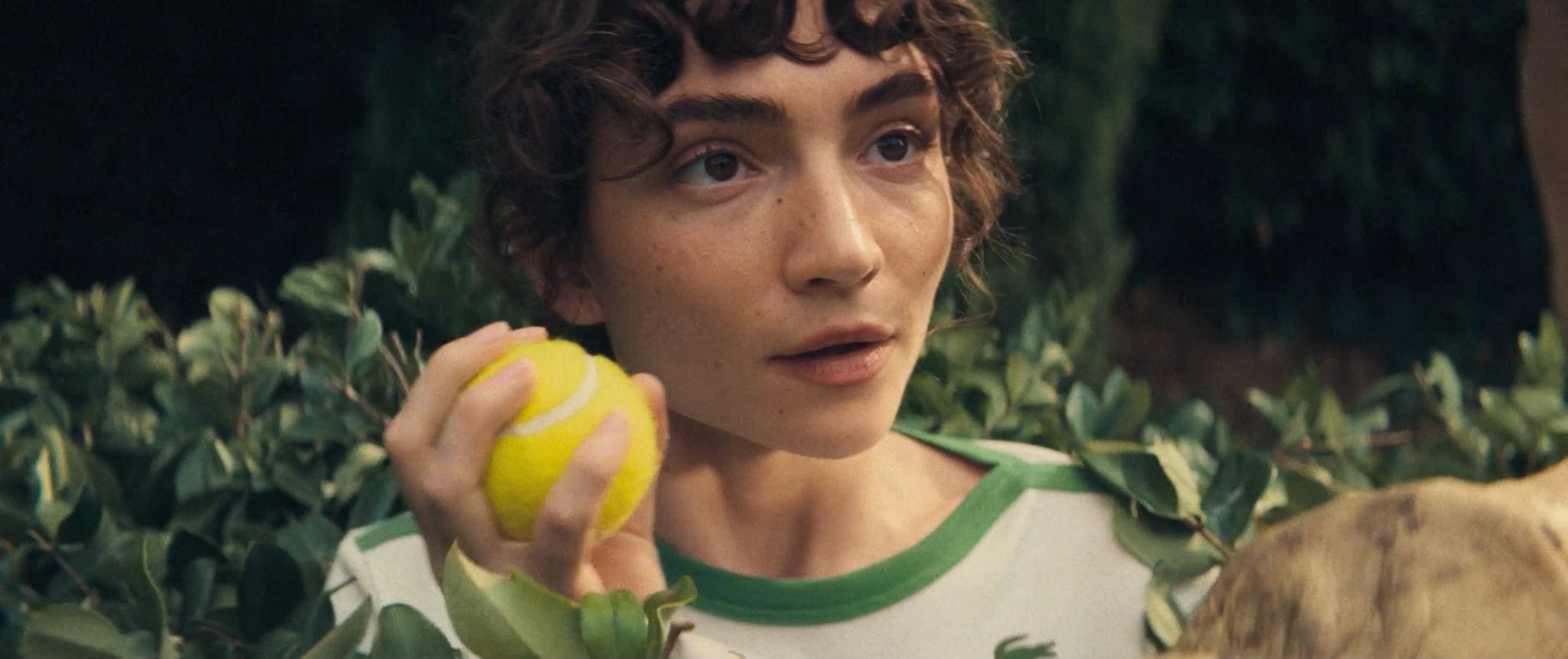 Close-up of a person with short curly hair holding a bright yellow tennis ball while surrounded by lush green foliage.
