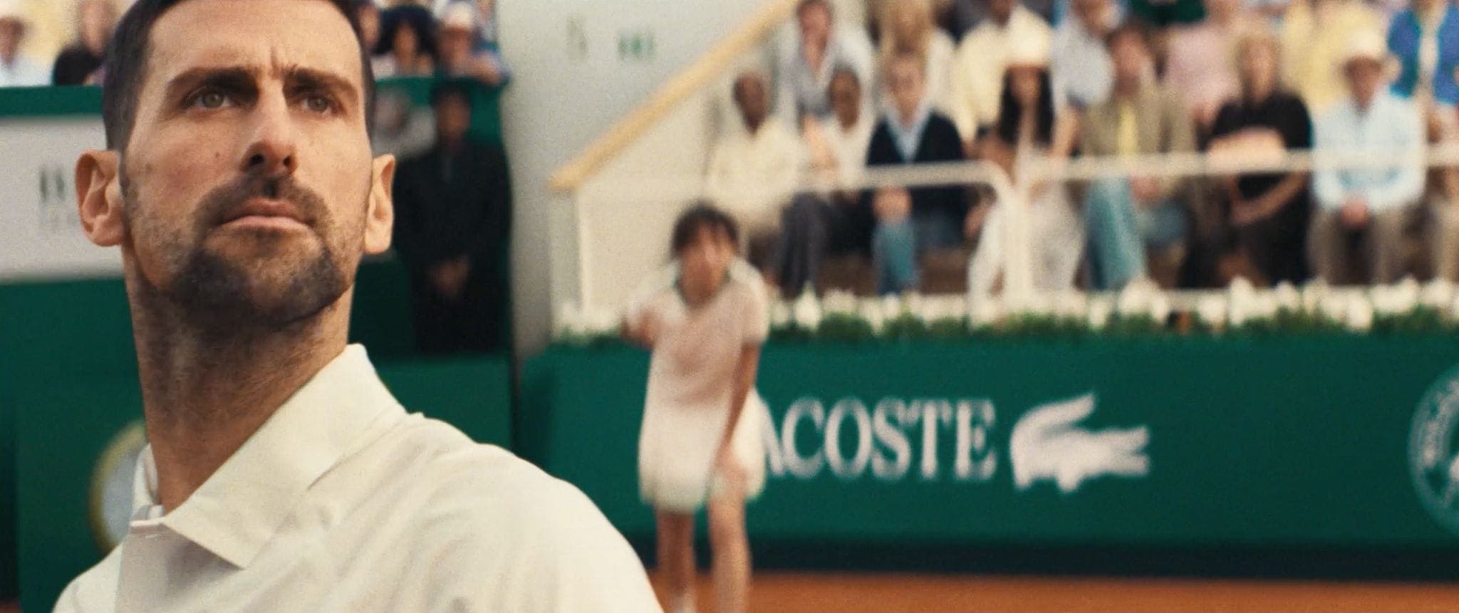 Novak Djokovic looking upward in a white polo shirt on a clay tennis court with Lacoste branding in the background.