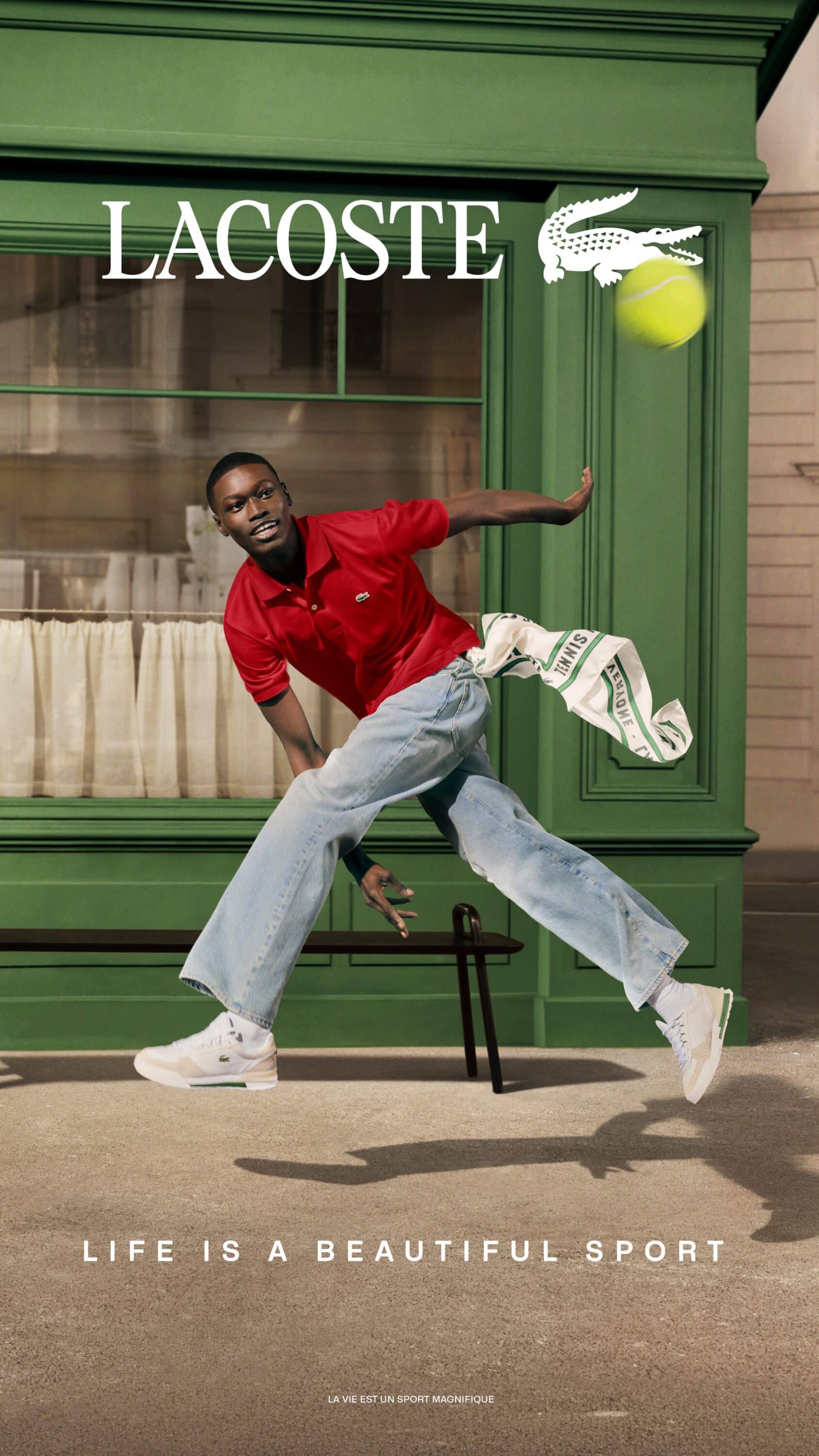 A man in a red polo shirt and light blue denim jeans leaping mid-air outside a green storefront next to a floating tennis ball.