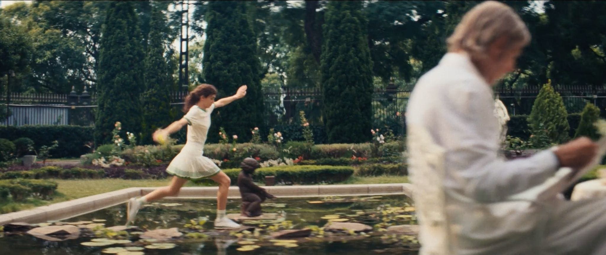 A woman in a white tennis dress leaps across stones in a garden pond while an out-of-focus man reads in the foreground.