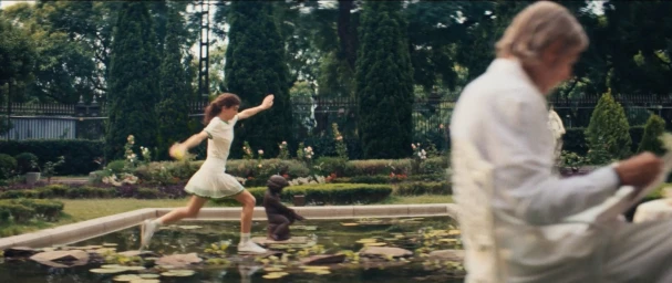 A woman in a white tennis dress leaps across stones in a garden pond while an out-of-focus man reads in the foreground.