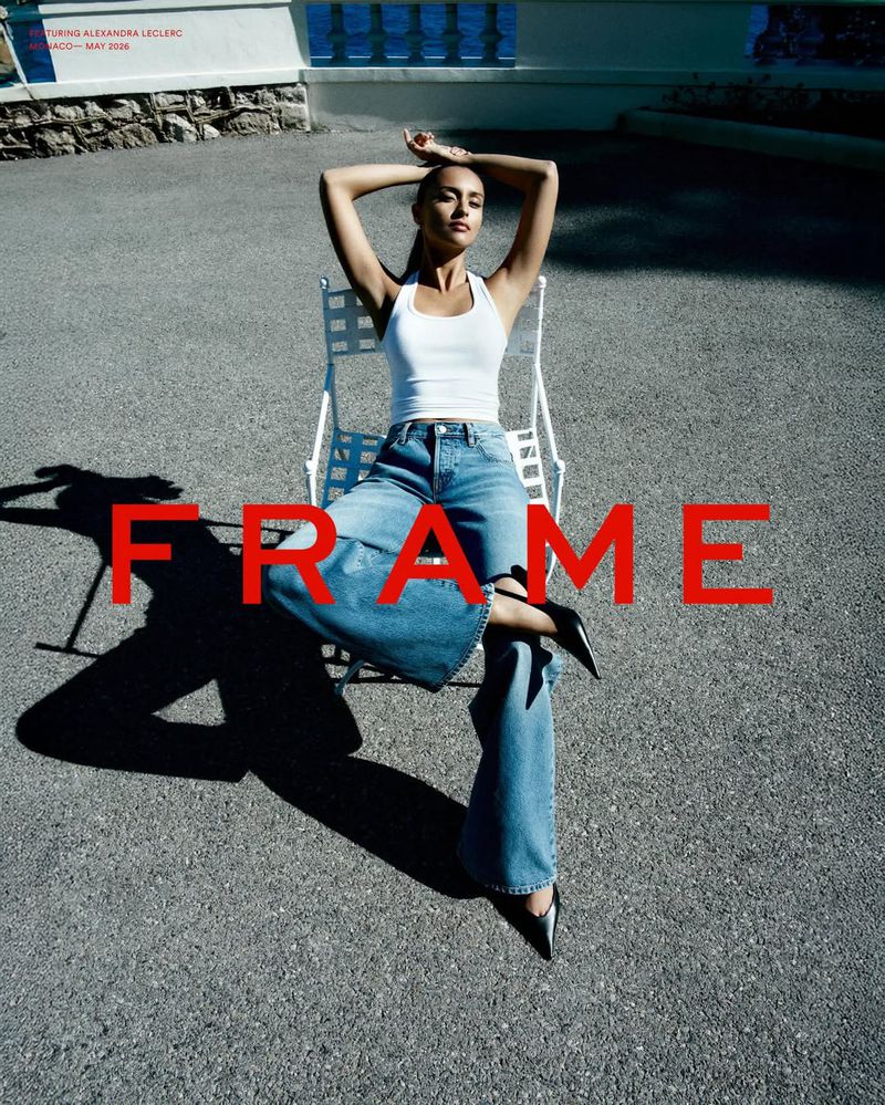 Alexandra Leclerc wearing a white tank top and wide-leg denim jeans seated on a white chair in direct sunlight.