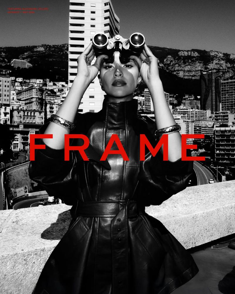 Monochrome portrait of a woman in a black leather trench coat holding binoculars on a rooftop overlooking a city.