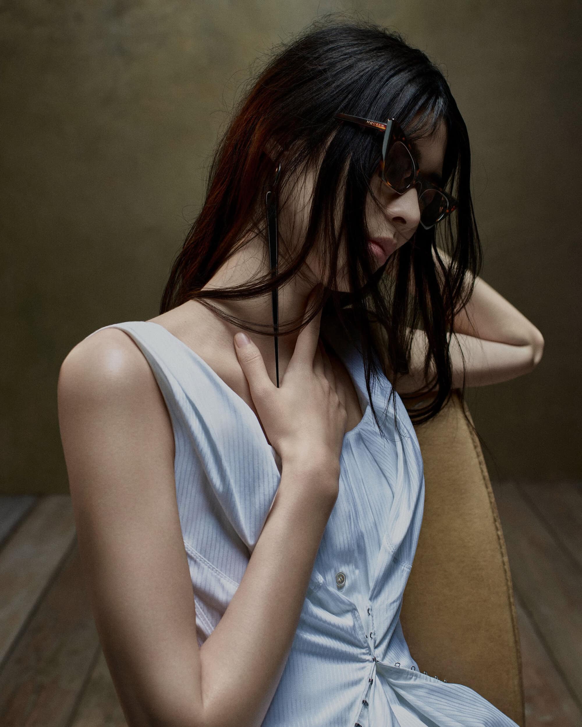 Model wearing dark tortoiseshell sunglasses and a sleeveless light dress while resting against a chair.