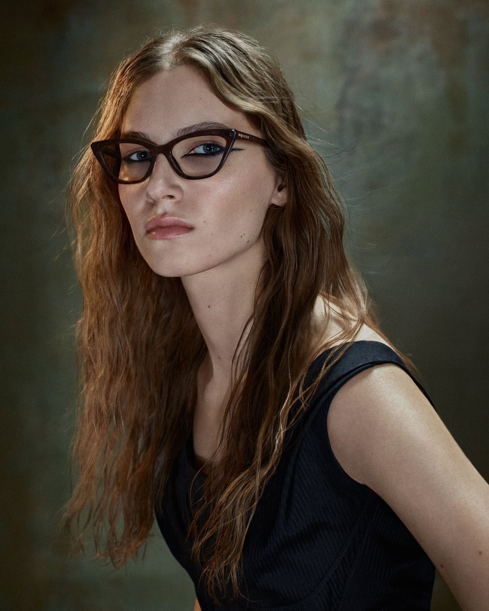Young woman wearing dark angular cat-eye glasses and a black ribbed top against a mottled olive textured backdrop.