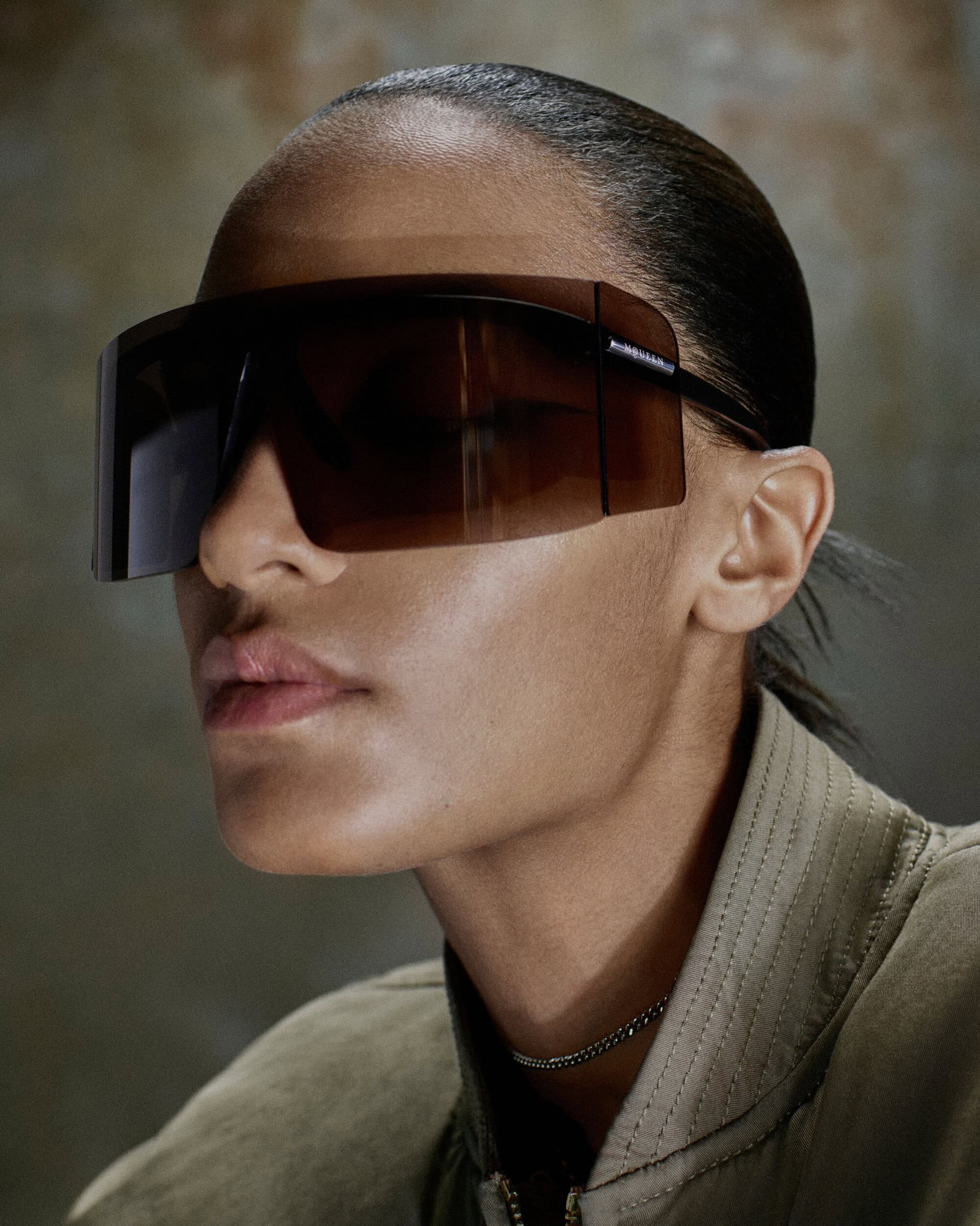 Model wearing oversized dark shield sunglasses and an olive green bomber jacket in a close-up profile portrait.