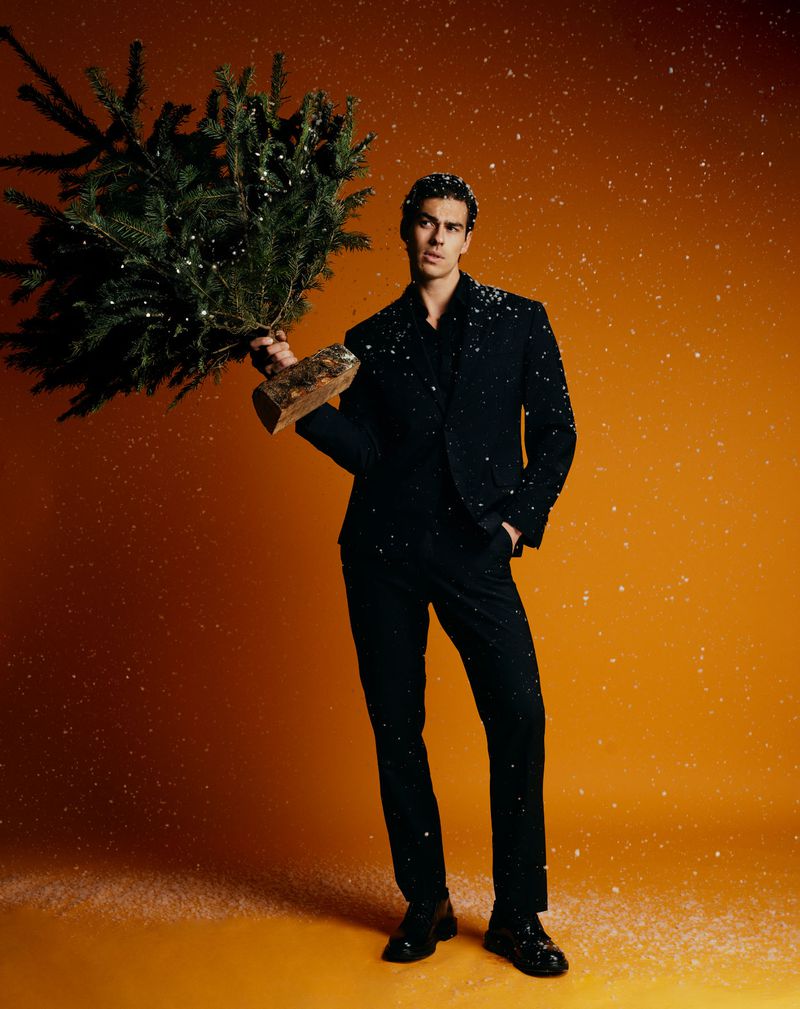Full-length portrait of a man in a black suit holding a small Christmas tree on an orange background with faux snow.
