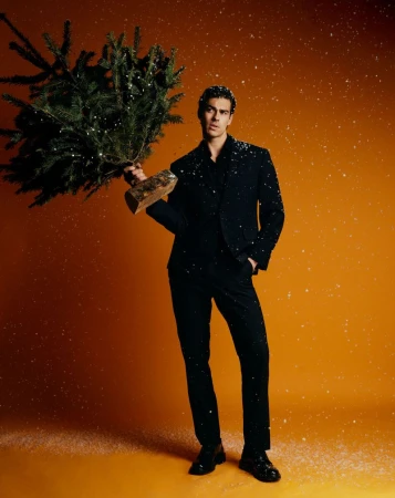 Full-length portrait of a man in a black suit holding a small Christmas tree on an orange background with faux snow.