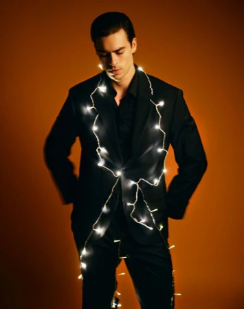 Young adult man in a black suit jacket and shirt adorned with white string lights in a studio setting with an orange backdrop.