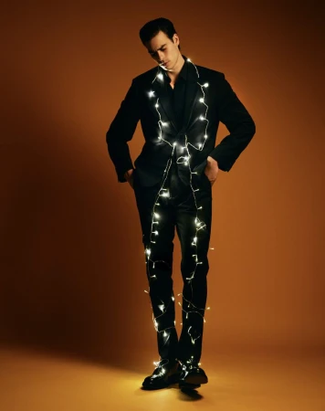Full-length studio shot of a young adult male in a black suit adorned with string lights against an orange background, fashion-forward and unique.