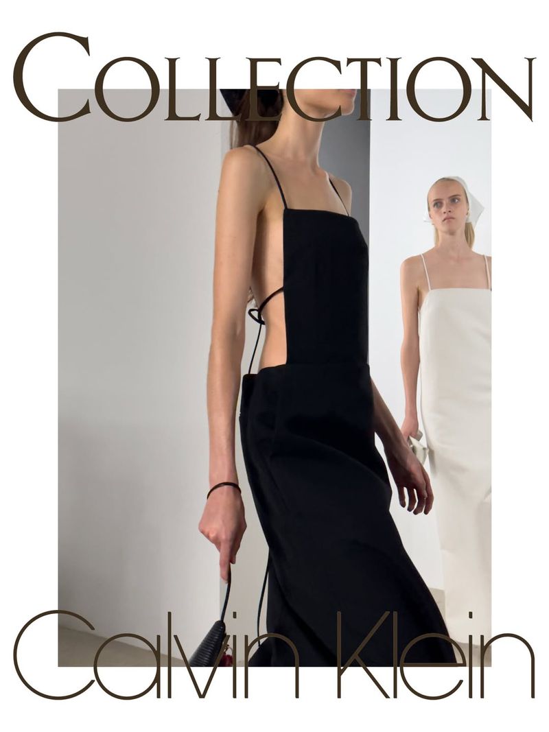 Model in a black slip dress walking past another model in white against a grey and white studio backdrop