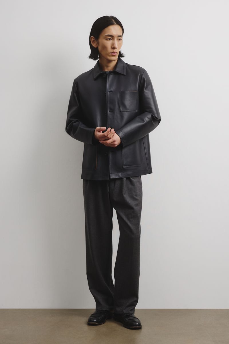 Full-length shot of a male model with dark hair in a black leather-look collared shirt and loose pants, posing against a white wall.