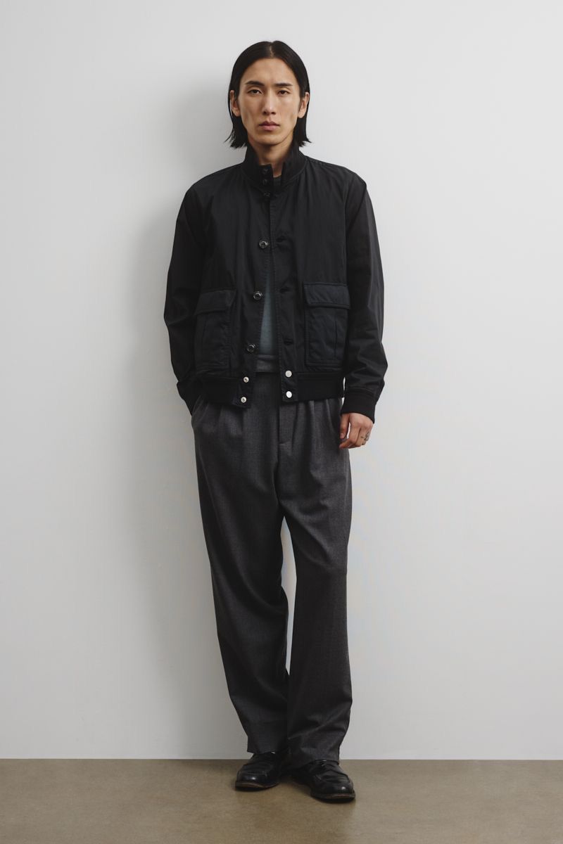 An adult male poses in a black jacket and grey slacks against a neutral wall, captured in a full shot with soft studio lighting.