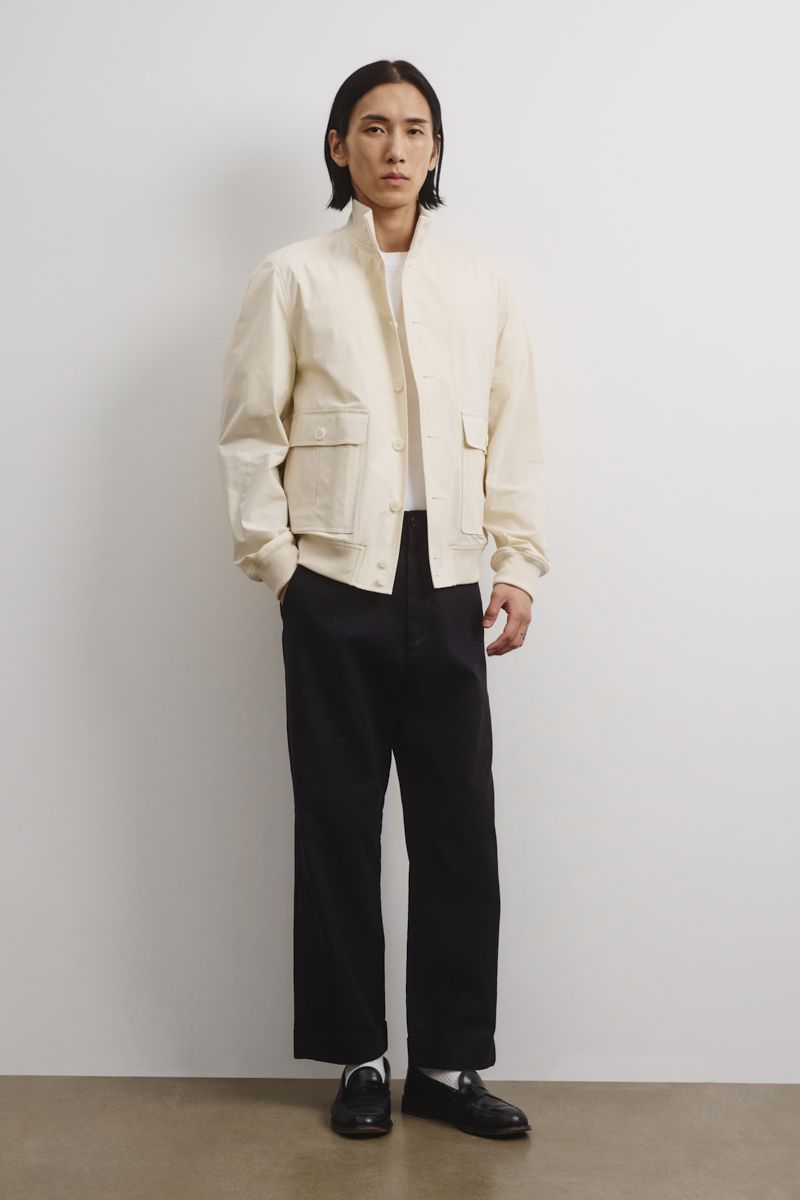 A full shot showcases a stylish adult male model with dark hair, posing in a light-colored jacket, dark pants, and loafers against a white wall.