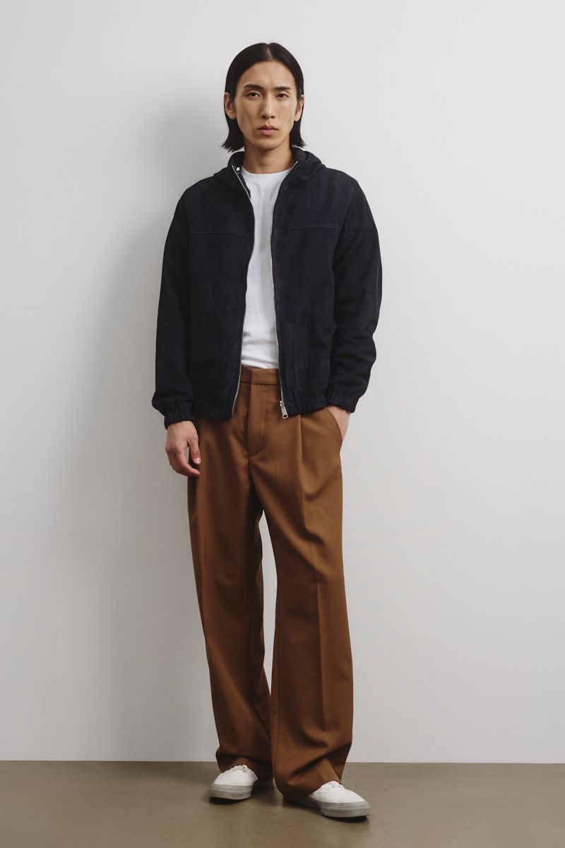 A slender adult man is posing in a navy zip jacket, white t-shirt, and brown pleated slacks against a white seamless backdrop.