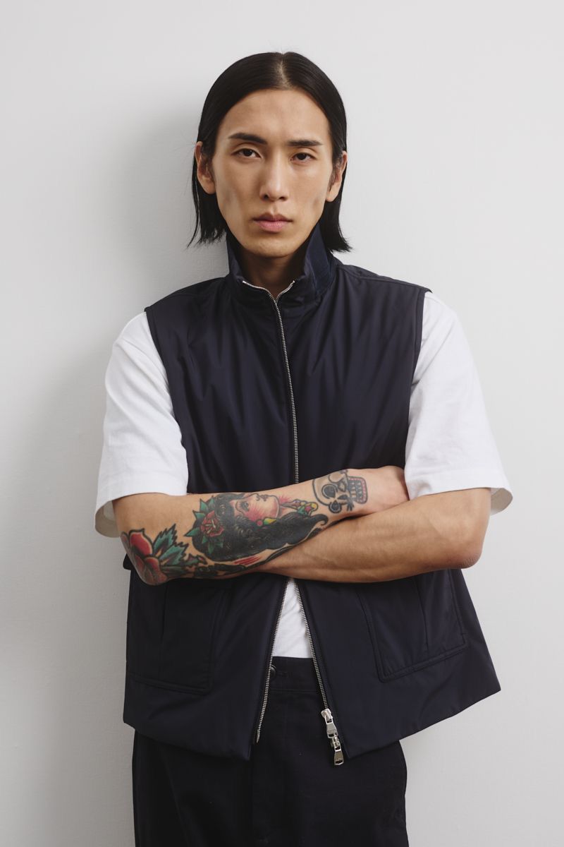 Portrait of a young Asian man with long dark hair wearing a navy vest, white t-shirt, and black pants with arms crossed against a white wall.