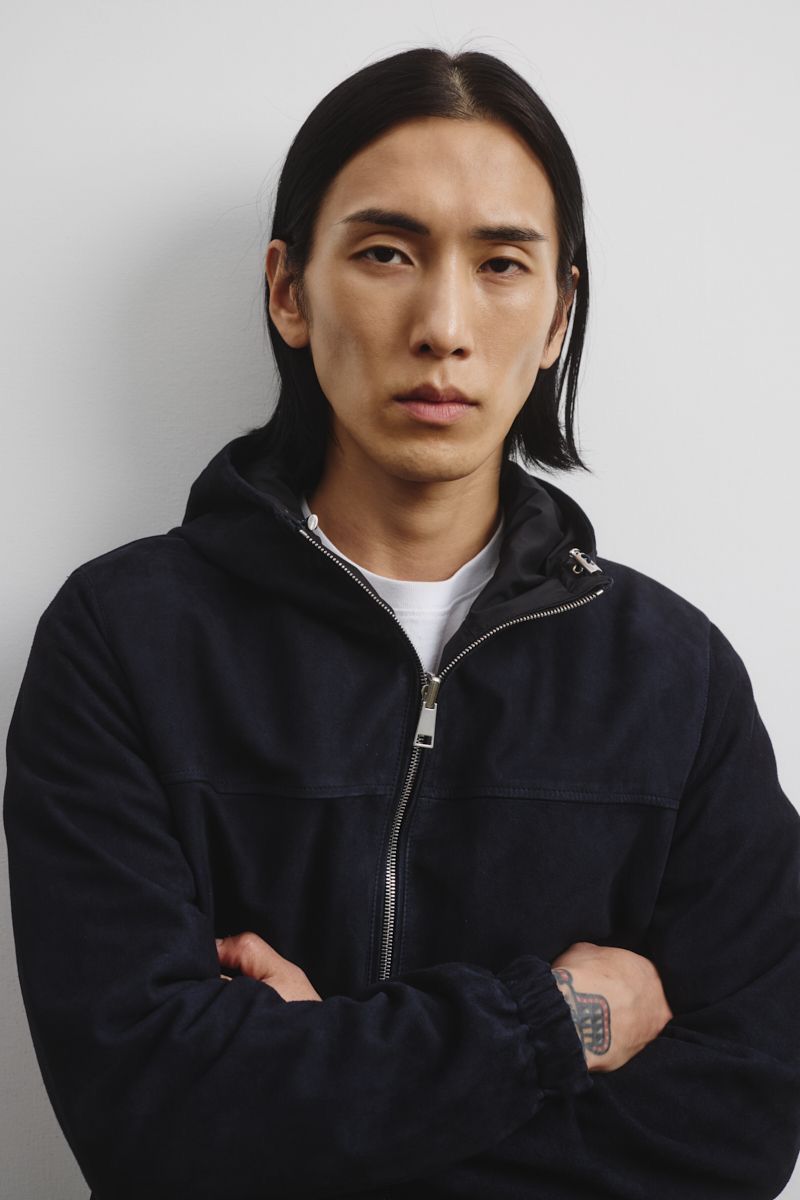 Portrait of an adult male wearing a hooded jacket with his arms crossed against a plain white background in a photo studio setting.