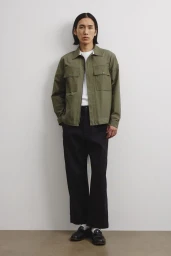 Model wearing an olive green utility jacket over a white t-shirt and dark trousers against a white seamless background