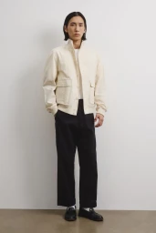 Model Hyuk Kim wearing a cream jacket, black wide-leg trousers, and black loafers standing against a pale studio wall.
