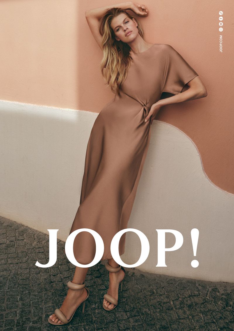 Kim Riekenberg in a taupe satin midi dress and high heels leaning against a peach and white painted wall.