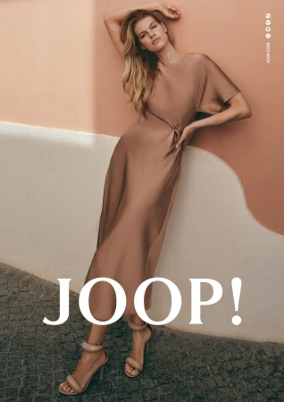 Kim Riekenberg in a taupe satin midi dress and high heels leaning against a peach and white painted wall.