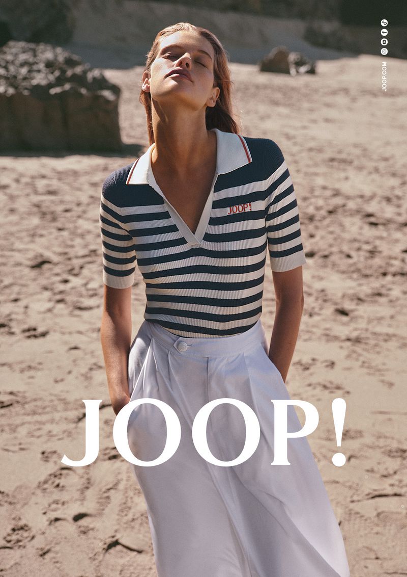 Model Kim Riekenberg wearing a navy striped knit polo shirt and white trousers on a sunny sandy beach.