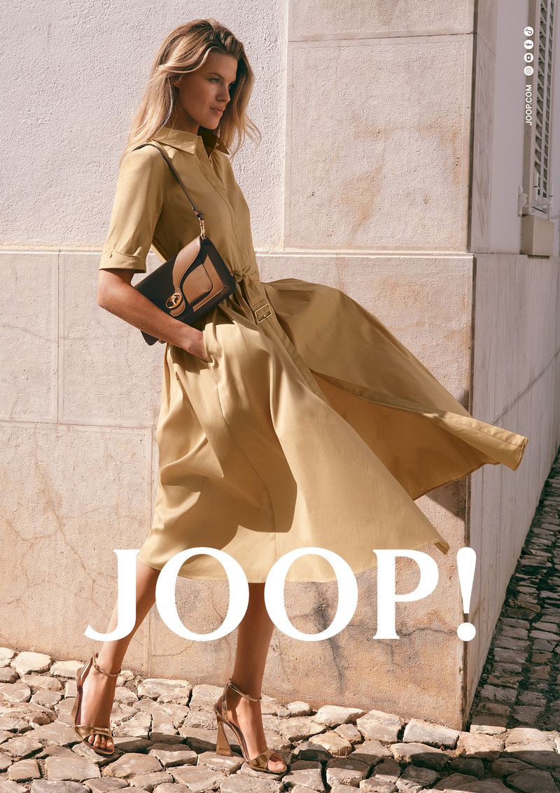 Model Kim Riekenberg in a flowing beige midi shirt dress and metallic heels walking along a cobblestone street for Joop.