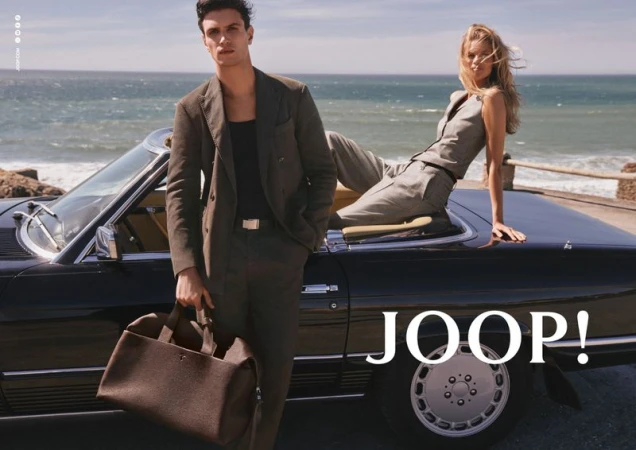Man standing beside a classic car holding a duffel bag with a woman seated on the trunk against a coastal background