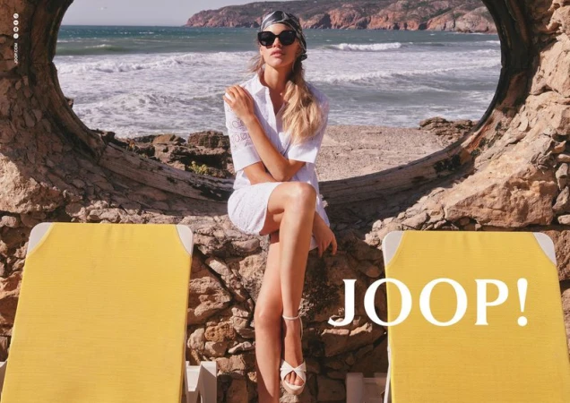 Kim Riekenberg sitting in a stone frame above the ocean in a white dress, sunglasses, and headscarf with yellow lounge chairs.