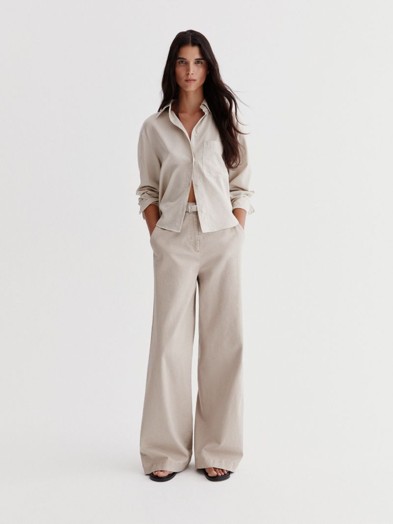 Model Paula Soares poses in a beige collared shirt and matching wide-leg trousers against a neutral studio backdrop.