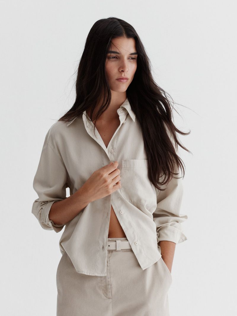 Model Paula Soares wearing an unbuttoned beige long-sleeve shirt and matching trousers against a white seamless studio backdrop.