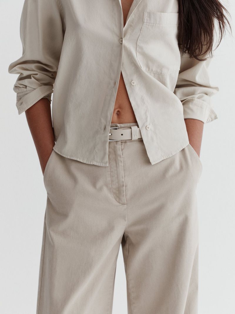 Close-up crop of a model wearing high-waisted beige trousers, a matching belt, and an unbuttoned button-down shirt.