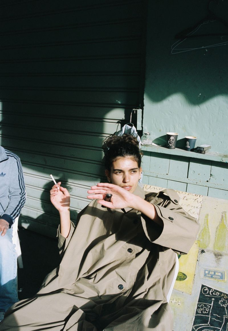 A young woman in a tan trench coat sits and smokes outside with a companion, with neutral wall art and coffee cups in the background.