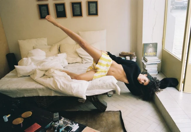 Model Sun Mizrahi reclines backward on a bed with one leg raised, wearing yellow striped shorts and a black sweater.