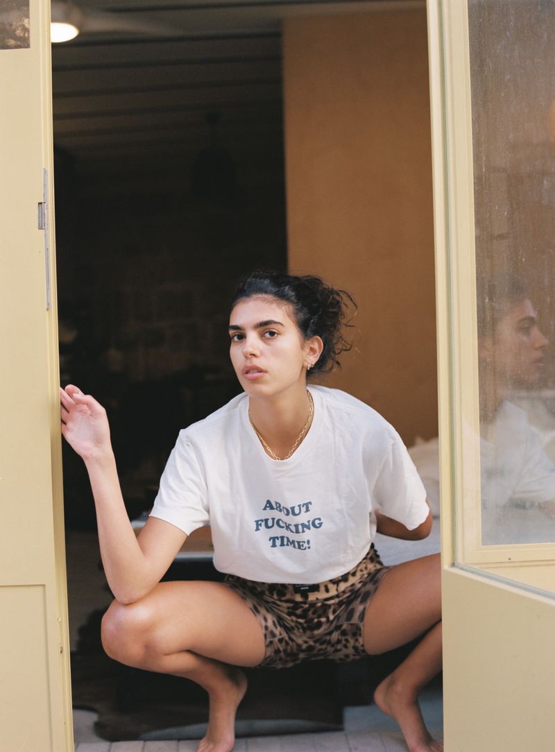Adult female poses in doorway wearing T-shirt with text and animal-print shorts. Casual, candid portrait with eye-level angle.