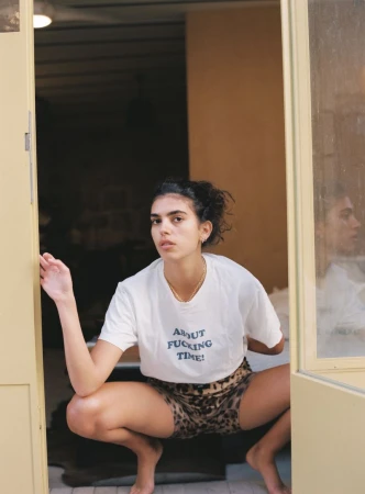 Adult female poses in doorway wearing T-shirt with text and animal-print shorts. Casual, candid portrait with eye-level angle.