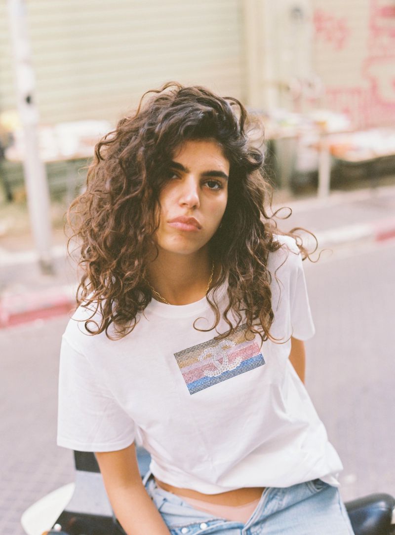 Model Sun Mizrahi with long curly brown hair wearing a white graphic t-shirt and jeans outdoors on a city street.